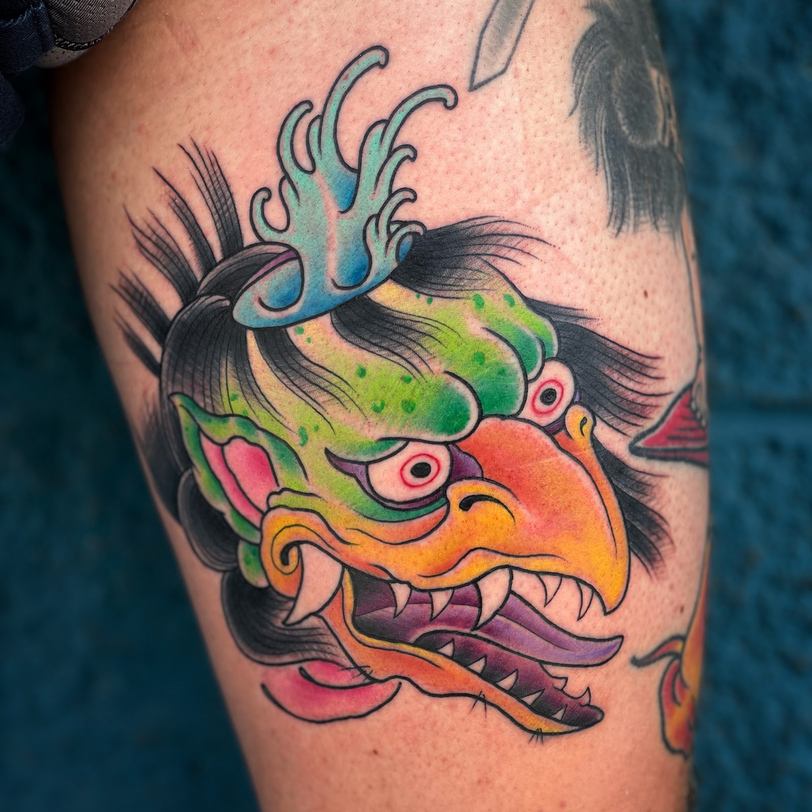 Japanese-style tengu head tattoo in bright green and orange, photographed against the shop's teal wall